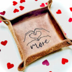 Small leather tray surrounded by love hearts. With an image of some hands held in a heart shape and the word more engraved on the centre of the tray